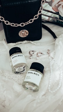 A black leather handbag with a gold chain sits on a delicate lace fabric. Nearby, there are two small bottles labeled 'DIALECT.' Earrings with pearl accents and a pearl necklace are also arranged on the lace.