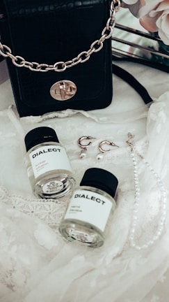 A black leather handbag with a gold chain sits on a delicate lace fabric. Nearby, there are two small bottles labeled 'DIALECT.' Earrings with pearl accents and a pearl necklace are also arranged on the lace.