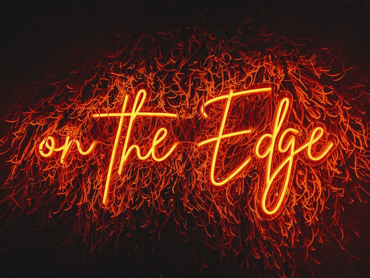 A vibrant neon sign displaying the phrase 'on the Edge' in cursive lettering against a dark background. The letters are illuminated in a bright orange color, creating a striking contrast with the surrounding darkness. The neon light is surrounded by abstract, flame-like patterns, adding a dynamic and edgy feel to the composition.
