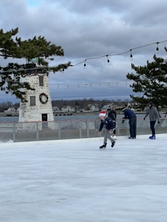 A group of people ice skating on a rink near a stone tower decorated with a wreath. The sky is overcast, and strings of lights hang above the rink. Evergreen trees frame the scene, and there are houses in the distance.