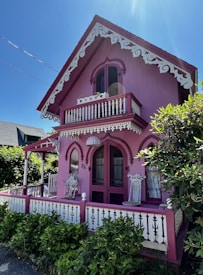 A charming, small, purple-pink house with intricate white trimmings and lace-like designs under the eaves. It features Gothic-style arched windows and a decorative balustrade on the balcony. The house is surrounded by lush green plants and the sky is bright and clear.