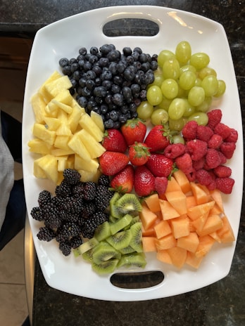 A platter filled with a variety of fresh fruits including blueberries, green grapes, raspberries, strawberries, pineapple chunks, blackberries, kiwi slices, and cantaloupe cubes, all arranged neatly on a white tray with handles.