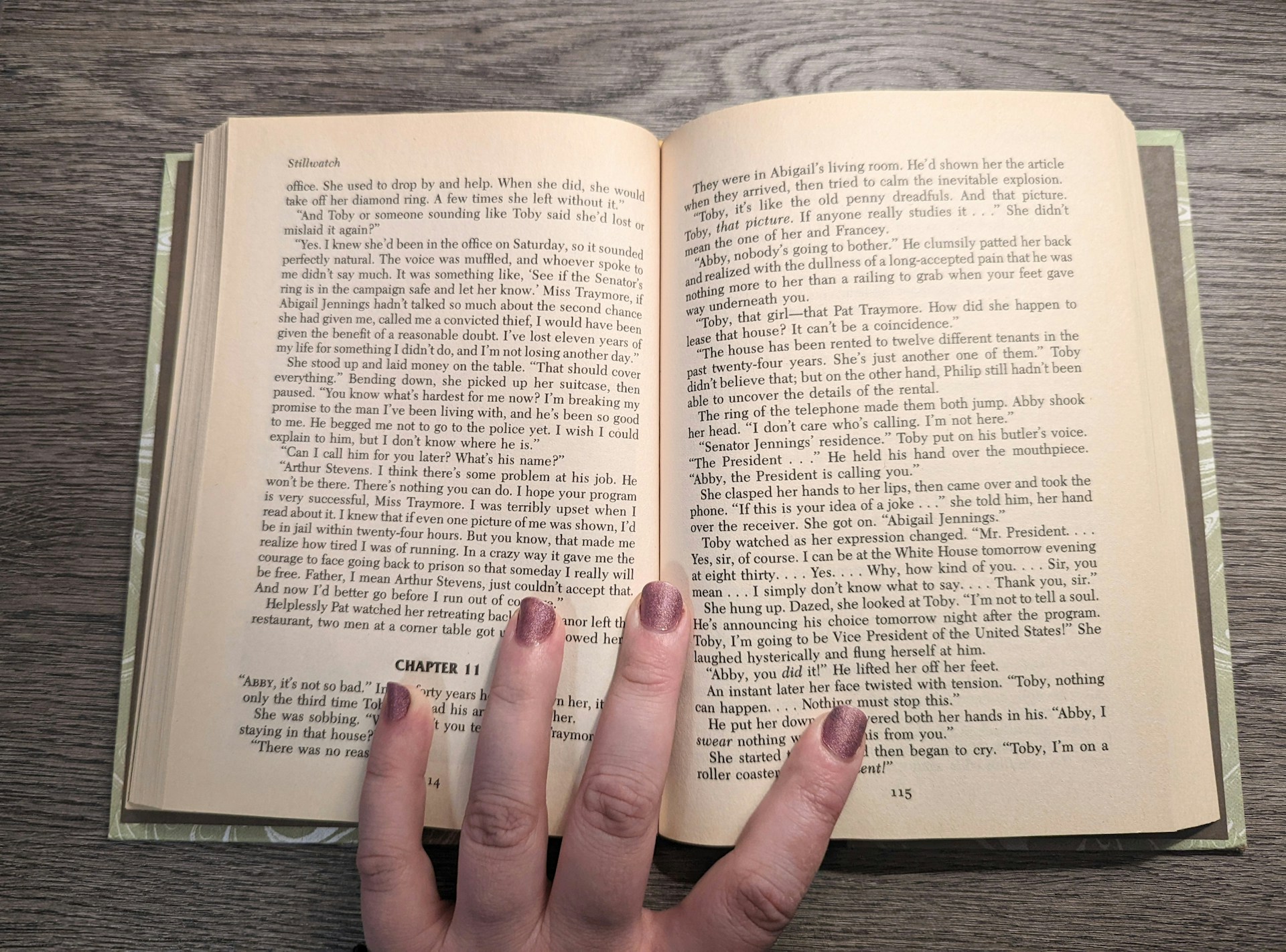 An open book rests on a wooden surface, revealing two pages filled with text. The book is held open by a hand with painted nails, which is positioned at the bottom center of the pages. The text appears to be a narrative or novel, with distinct paragraph formatting and a chapter heading labeled 'CHAPTER 11.' The fingernails are painted in a shimmering, neutral shade.