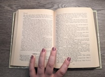 An open book rests on a wooden surface, revealing two pages filled with text. The book is held open by a hand with painted nails, which is positioned at the bottom center of the pages. The text appears to be a narrative or novel, with distinct paragraph formatting and a chapter heading labeled 'CHAPTER 11.' The fingernails are painted in a shimmering, neutral shade.
