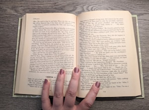 An open book rests on a wooden surface, revealing two pages filled with text. The book is held open by a hand with painted nails, which is positioned at the bottom center of the pages. The text appears to be a narrative or novel, with distinct paragraph formatting and a chapter heading labeled 'CHAPTER 11.' The fingernails are painted in a shimmering, neutral shade.