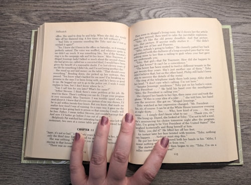 An open book rests on a wooden surface, revealing two pages filled with text. The book is held open by a hand with painted nails, which is positioned at the bottom center of the pages. The text appears to be a narrative or novel, with distinct paragraph formatting and a chapter heading labeled 'CHAPTER 11.' The fingernails are painted in a shimmering, neutral shade.