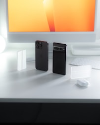 A modern workspace setup with two smartphones standing vertically on a clean white desk, next to a keyboard and a computer monitor displaying a bright orange and yellow gradient. Small white accessories are scattered around the desk, accentuating the organized and tech-savvy environment.