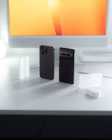 A modern workspace setup with two smartphones standing vertically on a clean white desk, next to a keyboard and a computer monitor displaying a bright orange and yellow gradient. Small white accessories are scattered around the desk, accentuating the organized and tech-savvy environment.