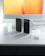A modern workspace setup with two smartphones standing vertically on a clean white desk, next to a keyboard and a computer monitor displaying a bright orange and yellow gradient. Small white accessories are scattered around the desk, accentuating the organized and tech-savvy environment.