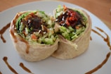 Two neatly cut sections of a wrap filled with fresh vegetables, drizzled with a dark sauce. The filling includes green lettuce, red bell pepper, and possibly bits of carrot or cabbage, adding a vibrant mix of colors and fresh appearance. The wrap is placed on a white plate, accompanied by decorative swirls of sauce.