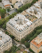 Aerial view of an urban area featuring a cluster of apartment buildings with gray rooftops and traditional European architectural style. The streets surrounding the buildings are lined with trees, and a few cars can be seen parked and moving along the roads. Crosswalks and some greenery are also visible in the vicinity.