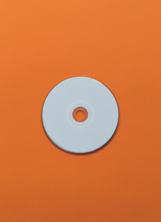 A single round, reflective disc, possibly a CD or DVD, lies against a bright orange textured background. The disc's center hole and shiny surface are prominent features within the composition.
