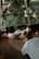 A person is lying on a woven mat inside a room with large windows, which reveal a lush, green forest outside. The person is illuminated by soft, natural light filtering through the window, creating a serene atmosphere. The room features wooden flooring and a bed with white sheets.