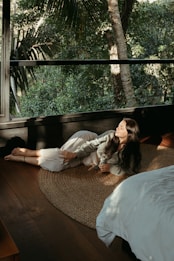 A person is lying on a woven mat inside a room with large windows, which reveal a lush, green forest outside. The person is illuminated by soft, natural light filtering through the window, creating a serene atmosphere. The room features wooden flooring and a bed with white sheets.