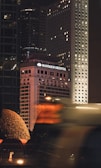 A bustling urban scene at night featuring the illuminated facade of the Mandarin Oriental hotel among a cluster of high-rise buildings. The buildings are adorned with numerous lit windows, and a red hue casts an ambient glow. In the foreground, blurred motion suggests the presence of a vehicle or pedestrian traffic, adding a dynamic element to the cityscape.