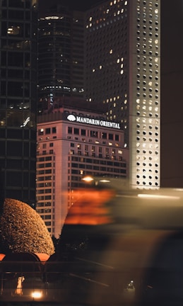 A bustling urban scene at night featuring the illuminated facade of the Mandarin Oriental hotel among a cluster of high-rise buildings. The buildings are adorned with numerous lit windows, and a red hue casts an ambient glow. In the foreground, blurred motion suggests the presence of a vehicle or pedestrian traffic, adding a dynamic element to the cityscape.