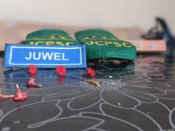 A name tag with the text 'JUWEL' in white letters on a blue background is placed on a dark floral-patterned surface. Behind it are green epaulettes with yellow text. Small red flowers are scattered on the surface.