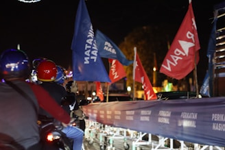 A group of people are riding motorcycles adorned with various flags, including blue and red ones. The background is dimly lit with streetlights visible, and a blue banner with white text can be seen over barricades. The setting appears to be a nighttime event or rally.