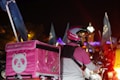 A food delivery person is on a motorcycle at night, wearing a pink and white helmet and jacket. The delivery box on the back of the motorcycle is also pink, featuring a panda logo. Blue flags and lights can be seen in the background, indicating a lively atmosphere.