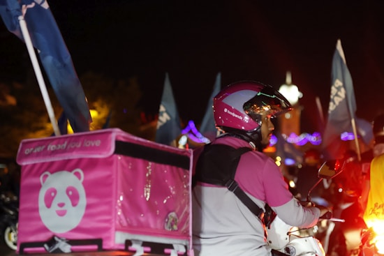 A food delivery person is on a motorcycle at night, wearing a pink and white helmet and jacket. The delivery box on the back of the motorcycle is also pink, featuring a panda logo. Blue flags and lights can be seen in the background, indicating a lively atmosphere.