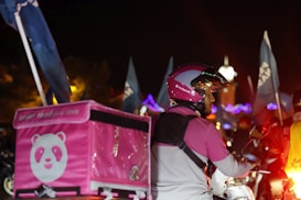 A food delivery person is on a motorcycle at night, wearing a pink and white helmet and jacket. The delivery box on the back of the motorcycle is also pink, featuring a panda logo. Blue flags and lights can be seen in the background, indicating a lively atmosphere.