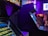 A dimly lit arcade with several classic arcade game machines in view. Neon lights glow, casting a purple and blue hue over the environment. Some text is visible on the game screens, with one machine having a prominent display of game features. The area seems to be bustling with activity with a few blurred figures visible in the background.