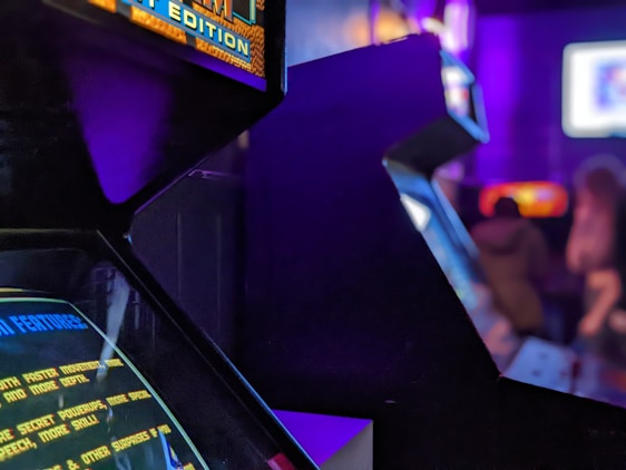 A dimly lit arcade with several classic arcade game machines in view. Neon lights glow, casting a purple and blue hue over the environment. Some text is visible on the game screens, with one machine having a prominent display of game features. The area seems to be bustling with activity with a few blurred figures visible in the background.