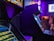 A dimly lit arcade with several classic arcade game machines in view. Neon lights glow, casting a purple and blue hue over the environment. Some text is visible on the game screens, with one machine having a prominent display of game features. The area seems to be bustling with activity with a few blurred figures visible in the background.