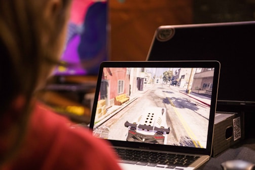 A person is playing a video game on a laptop, with the screen showing a car driving through a virtual city street. The surrounding environment includes several electronic devices and a table.