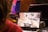 A person is playing a video game on a laptop, with the screen showing a car driving through a virtual city street. The surrounding environment includes several electronic devices and a table.
