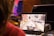A person is playing a video game on a laptop, with the screen showing a car driving through a virtual city street. The surrounding environment includes several electronic devices and a table.