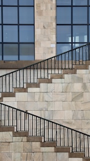 A stone wall with a series of ascending and descending staircases featuring dark metal railings. The wall is composed of rectangular stone tiles in light shades, and large grid-like windows are present at the top half of the frame.