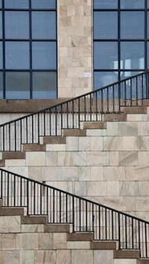 A stone wall with a series of ascending and descending staircases featuring dark metal railings. The wall is composed of rectangular stone tiles in light shades, and large grid-like windows are present at the top half of the frame.