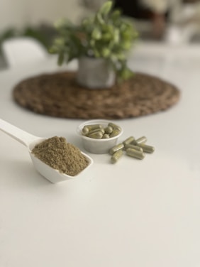 A white scoop filled with a fine greenish-brown powder is placed on a plain white surface. Nearby, there is a small white dish containing several green capsules. In the blurred background, a potted plant sits on a woven mat, adding a touch of greenery to the scene.