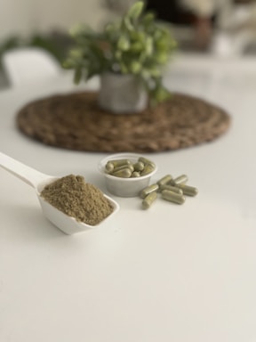A white scoop filled with a fine greenish-brown powder is placed on a plain white surface. Nearby, there is a small white dish containing several green capsules. In the blurred background, a potted plant sits on a woven mat, adding a touch of greenery to the scene.