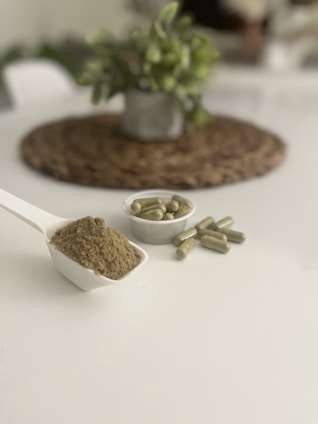A white scoop filled with a fine greenish-brown powder is placed on a plain white surface. Nearby, there is a small white dish containing several green capsules. In the blurred background, a potted plant sits on a woven mat, adding a touch of greenery to the scene.