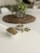 A white scoop filled with a fine greenish-brown powder is placed on a plain white surface. Nearby, there is a small white dish containing several green capsules. In the blurred background, a potted plant sits on a woven mat, adding a touch of greenery to the scene.
