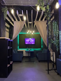 A cozy lounge area with two blue sofas facing a wall-mounted TV displaying a colorful image. Green plants hang from the ceiling and decorate the walls, which are draped with beige curtains. The ceiling features exposed beams and pendant lighting, and there are soft neon green accents along the edges.