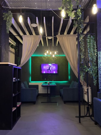 A cozy lounge area with two blue sofas facing a wall-mounted TV displaying a colorful image. Green plants hang from the ceiling and decorate the walls, which are draped with beige curtains. The ceiling features exposed beams and pendant lighting, and there are soft neon green accents along the edges.