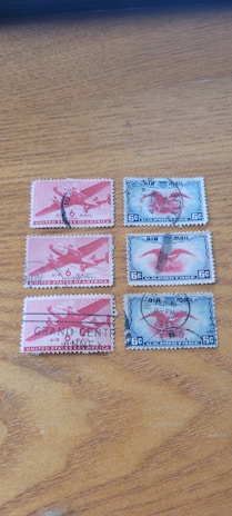Five vintage airmail postage stamps are arranged on a wooden surface. Three of the stamps are red and depict an airplane with '6 Cents United States Postage' inscribed. Each stamp has a postal cancellation mark. The other two stamps are blue, also showing an airplane, with '6 C US Postage' at the bottom. These stamps also bear postal cancellation marks.