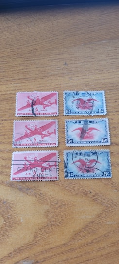 Five vintage airmail postage stamps are arranged on a wooden surface. Three of the stamps are red and depict an airplane with '6 Cents United States Postage' inscribed. Each stamp has a postal cancellation mark. The other two stamps are blue, also showing an airplane, with '6 C US Postage' at the bottom. These stamps also bear postal cancellation marks.