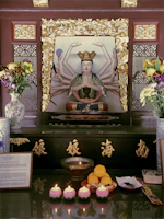 A detailed altar featuring a multi-armed statue, possibly a deity, surrounded by golden ornate frames. The altar is adorned with various offerings, including flowers in decorative vases, fruit, and lit candles. The setup conveys a sense of reverence and spirituality, enhanced by the elegant calligraphy and intricate designs.
