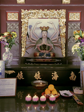 A detailed altar featuring a multi-armed statue, possibly a deity, surrounded by golden ornate frames. The altar is adorned with various offerings, including flowers in decorative vases, fruit, and lit candles. The setup conveys a sense of reverence and spirituality, enhanced by the elegant calligraphy and intricate designs.