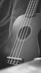 A close-up view of a guitar or ukulele resting on a fabric with visible strings and a circular soundhole. The image is in black and white, highlighting the texture of the fabric and the smooth surface of the instrument.