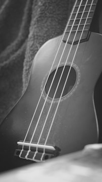 A close-up view of a guitar or ukulele resting on a fabric with visible strings and a circular soundhole. The image is in black and white, highlighting the texture of the fabric and the smooth surface of the instrument.