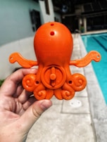 An orange plastic octopus toy is being held by a hand near a swimming pool. The toy appears to be a spray or water toy, with a circular mechanism visible on its back. The background shows a blurred poolside area, with water visible on the right.