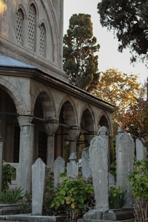 A historical cemetery with intricately carved tombstones surrounded by lush greenery and flowers. The tombstones are set against the backdrop of an ancient stone building with arched windows and decorative architectural details. Tall trees with autumn foliage are visible in the background.
