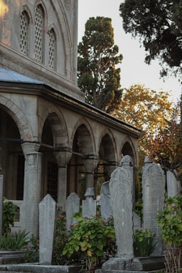 A historical cemetery with intricately carved tombstones surrounded by lush greenery and flowers. The tombstones are set against the backdrop of an ancient stone building with arched windows and decorative architectural details. Tall trees with autumn foliage are visible in the background.