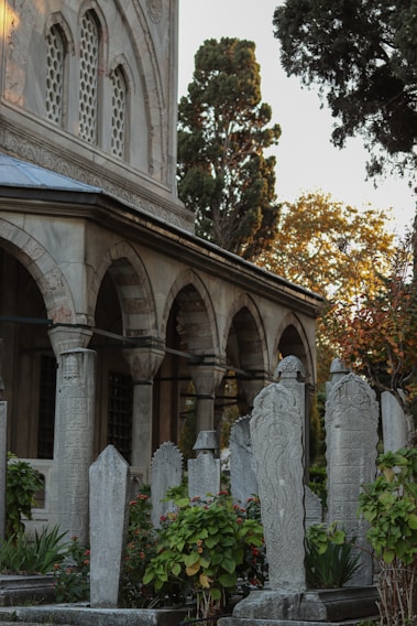 A historical cemetery with intricately carved tombstones surrounded by lush greenery and flowers. The tombstones are set against the backdrop of an ancient stone building with arched windows and decorative architectural details. Tall trees with autumn foliage are visible in the background.