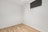 A small, empty room with light wooden flooring and plain white walls. A single high window with a dark blind lets in minimal light, creating a minimalist and sterile appearance.